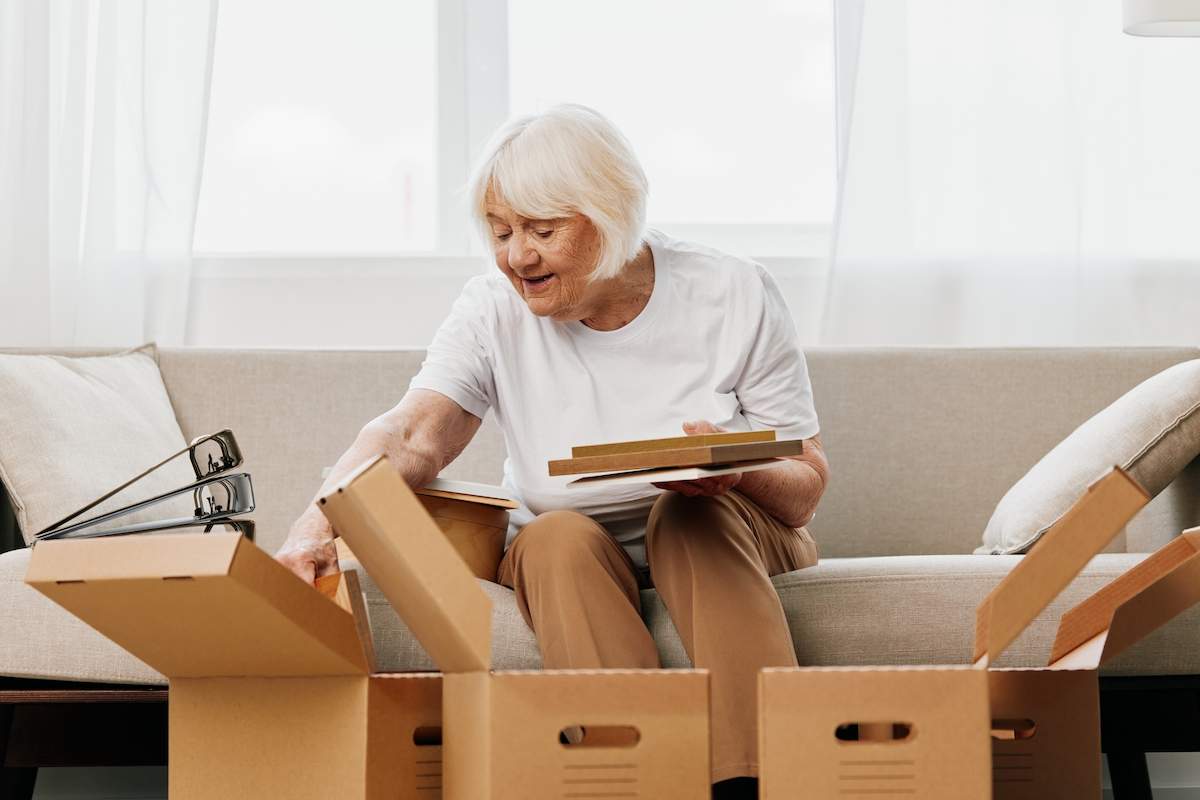Downsizing Isn't Downgrading: Tips for Upgrading Your Life elderly woman sits on a sofa at home with boxes. collecting things with memories albums with photos and photo frames moving to a new place cleaning things and a happy smile. Lifestyle retirement.